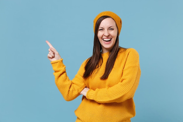 Laughing young brunette woman girl in yellow sweater hat posing isolated on blue background studio portrait. People sincere emotions lifestyle concept. Mock up copy space. Pointing index finger aside.