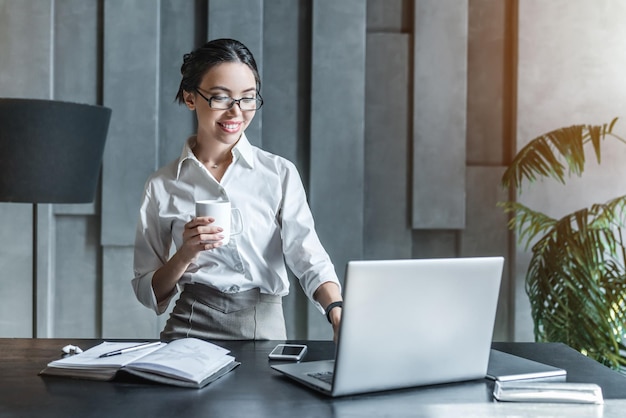 Laughing young female entrepreneur drinking cup of coffee and working on laptop in her home office