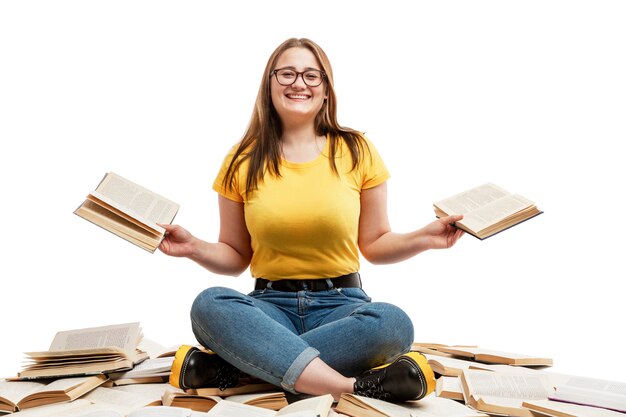 A laughing young girl in jeans and a blue T-shirt sits on a pile of opened books and holds books in her hands