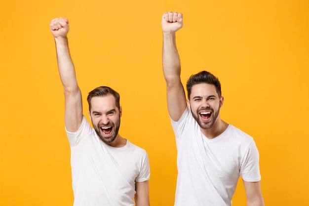 Laughing young men guys friends in white blank empty t-shirts posing isolated on yellow orange wall in . People lifestyle concept.  Rising hands up doing winner gesture.