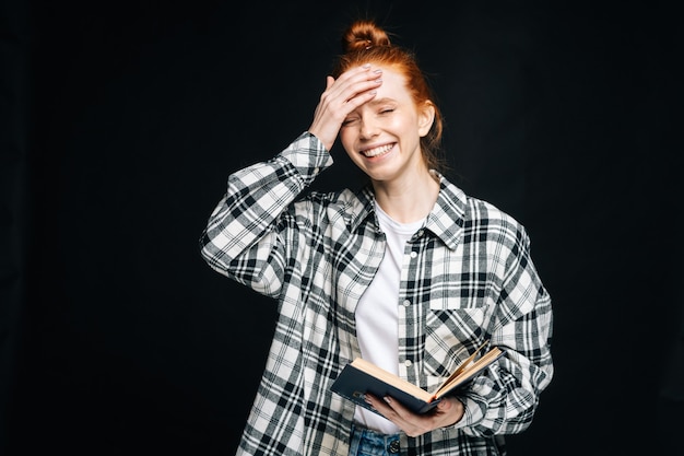 Laughing young woman college student holding opened book on black isolated background