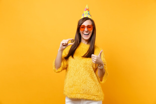 Laughing young woman in orange heart glasses, birthday hat showing thumb up holding bitcoin metal coin of golden color future currency isolated on yellow background. People sincere emotions lifestyle.