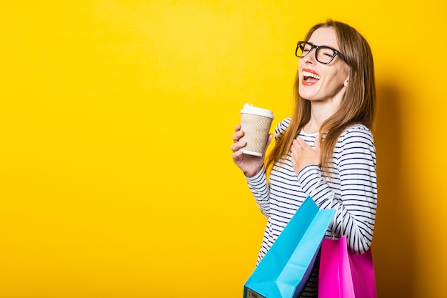 Laughing young woman rejoices holding a paper cup with coffee and bags on a yellow background