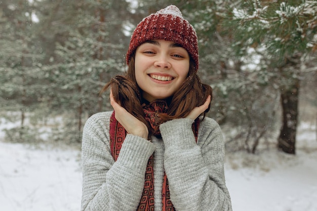 Laughing young woman in warm hat and scarf looking at camera in snowy winter forest