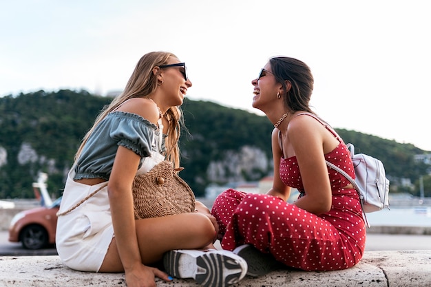 Laughing young women in summer outfit and sunglasses spending good time together while sitted