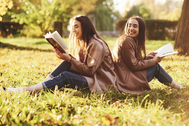 Laughinh brunette twin girls sitting back to back on the grass and having fun with legs slightly bent in knees, with brown books in hands, wearing casual coat in autumn park on blurry background.