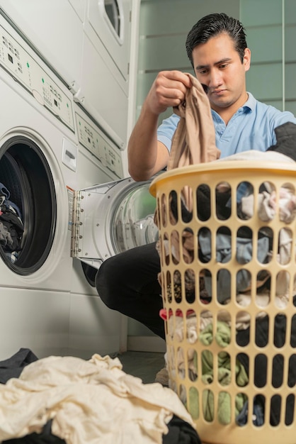 Laundry employee with basket full of laundry taking it outside to put it in the washing machine. Young adult male bending down to put clothes in the washing machine.