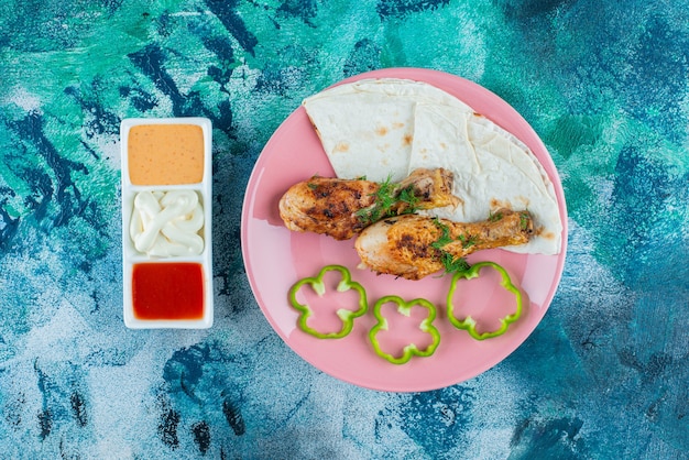 Lavash, baked drumsticks and pepper on a plate next to sauce bowls on the blue surface