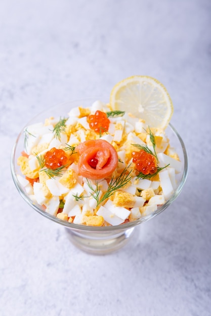 Layered salad with salted trout (salmon), caviar, eggs and vegetables. Traditional dish, one portion in a glass goblet on a gray background. Close-up.