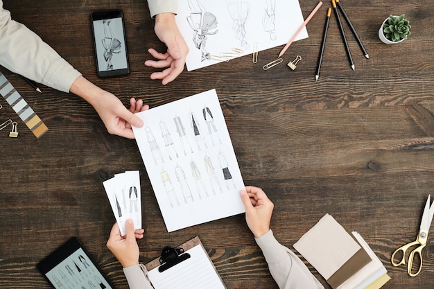 Photo layout of hands of creative designer passing paper with drawn sketches to his female colleague while both choosing fashion models over table