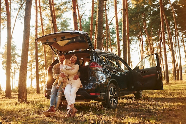 Leaning on the car Happy family of father mother and little daughter is in the forest