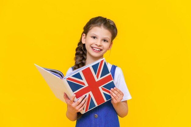 Learning English for children A little girl is holding a book with an English flag in her hands and smiling broadly on a yellow isolated background