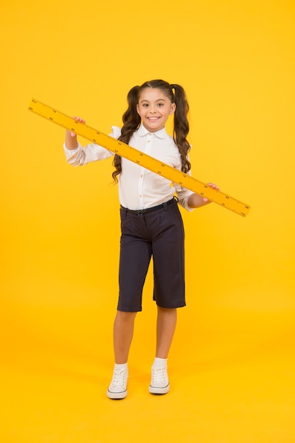 Learning geometry at school. Small school child holding measuring device on yellow background. Little girl preparing long wooden ruler for school lesson. Keep measuring and back to school.