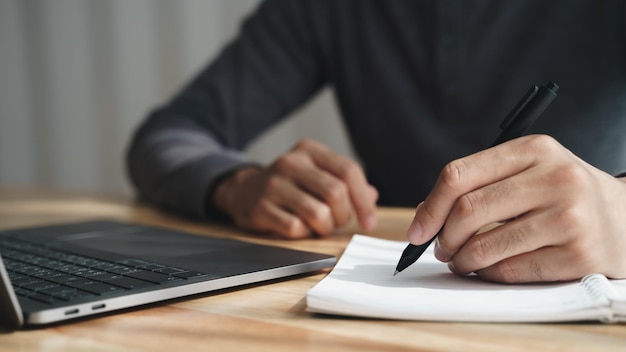 Left handed man writes in a notebook on the table with laptop computer