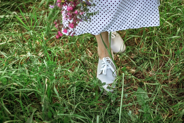 The legs of a beautiful girl in a long white dress and gray shoes are standing in the tall green grass. A girl holds a bouquet of purple wildflowers. Selective focus. Copy Space.