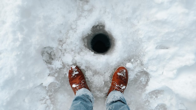 Legs of a fisherman in brown winter boots standing near a hole in the ice outdoors pov