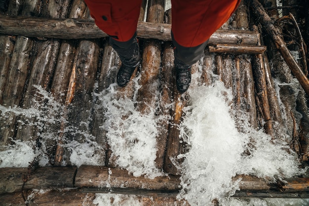 Legs of tourist in red trousers and trekking boots in water waves on log bridge across powerful mountain river. Traveler legs in water splashes across wooden bridge over mountain river with rapids.