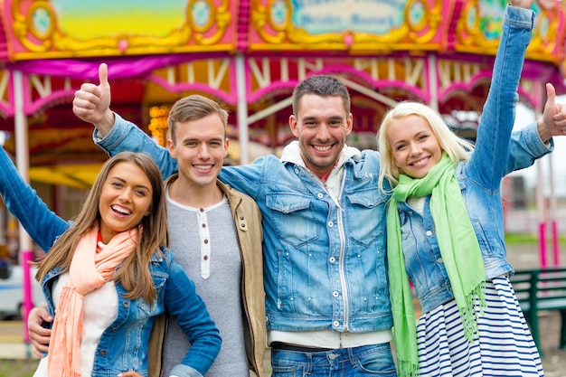 leisure, amusement park and friendship concept - group of smiling friends showing thumbs up with carousel on the back