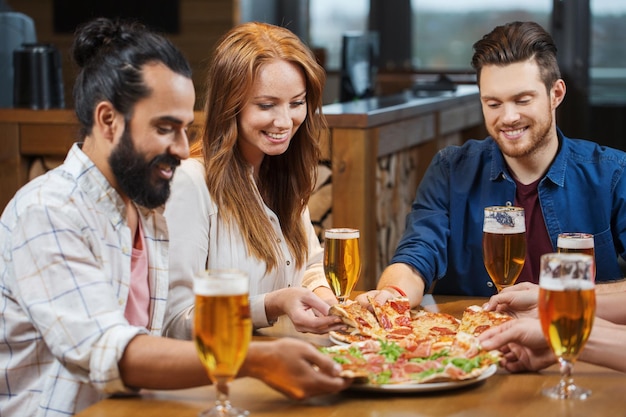 leisure, food and drinks, people and holidays concept - smiling friends eating pizza and drinking beer at restaurant or pub