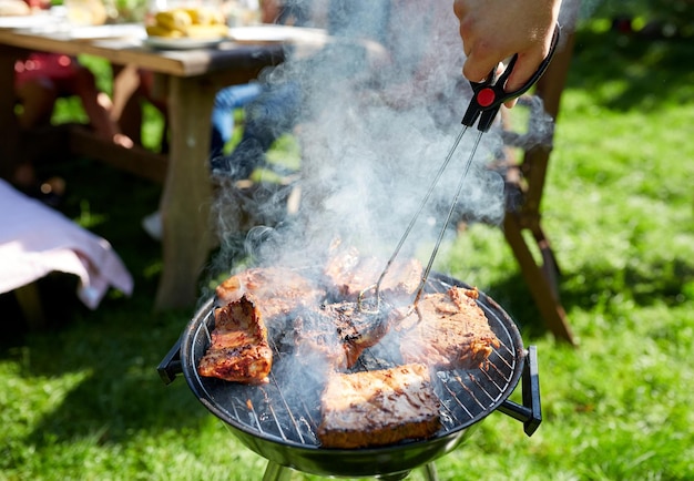 leisure, food, people and holidays concept - man cooking meat on barbecue grill for his friends at summer outdoor party