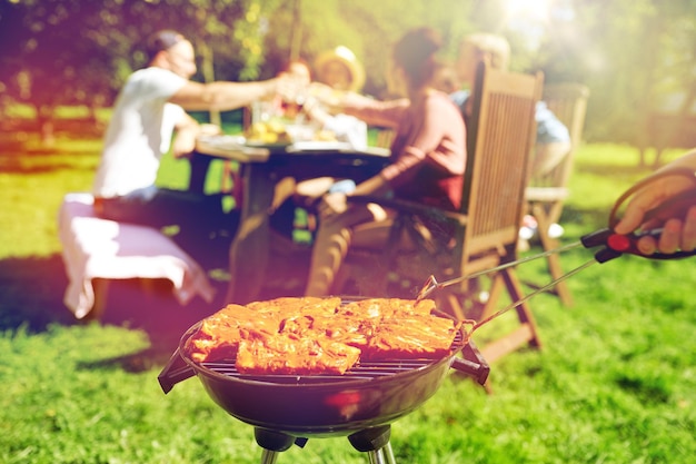 leisure, food, people and holidays concept - man cooking meat on barbecue grill for his friends at summer outdoor party