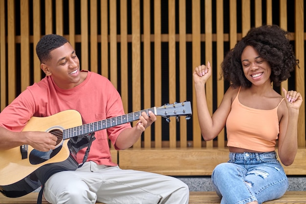 Leisure, guitar. Joyful young dark-skinned woman with raised arms and smiling man in casual clothes playing guitar sitting outdoors on fine day