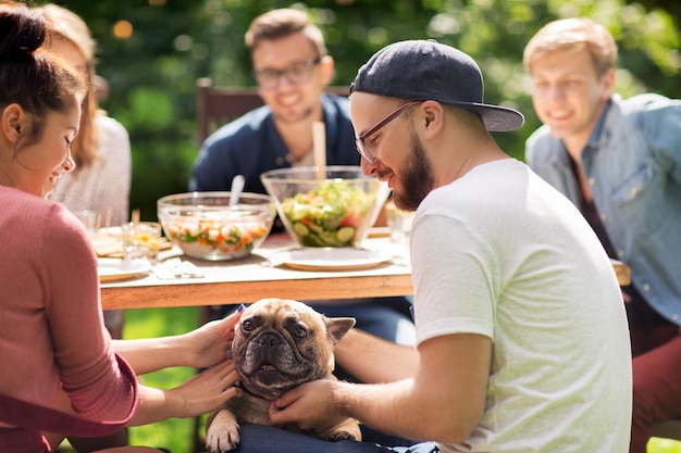 leisure, holidays, eating, people and food concept - happy friends with french bulldog dog having dinner at summer garden party