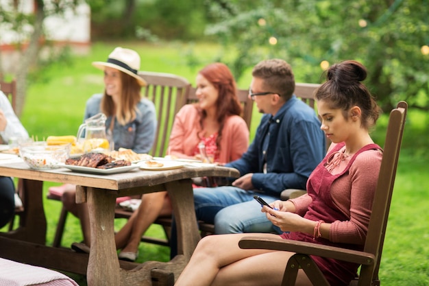 leisure, holidays, people and technology concept - young woman or teenage girl texting on smartphone and friends having dinner at summer garden party