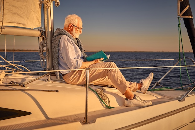 Leisure and relaxation senior man reading book while sitting on the side of sailboat or yacht deck