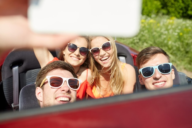 leisure, road trip, travel and people concept - happy friends driving in cabriolet taking selfie by smartphone outdoors