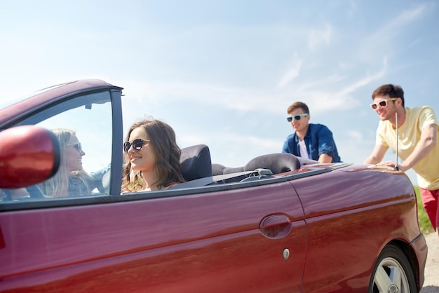 leisure, road trip, travel and people concept - happy friends pushing broken cabriolet car along country road