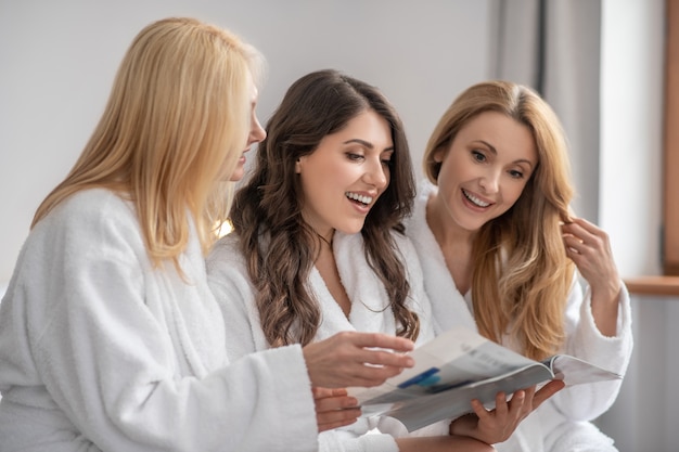 Photo leisure. smiling adult good looking women with long hair in white coats looking at magazine spending leisure time together in bright room