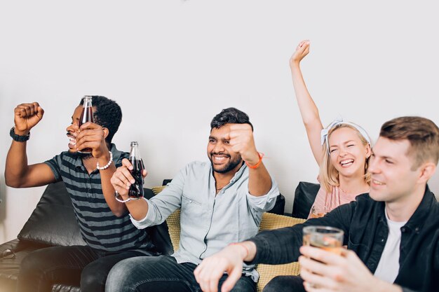 Photo leisure, sport, happiness concept. happy friends watching football match on tv sitting on couch with drinks, raising hands, shouting, celebrating goal, rejoicing victory of favorite team.