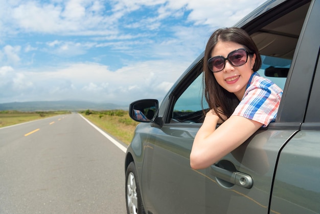 leisurely female tourist wearing sunglasses sitting on travel car and face to camera happily enjoying road trip at summer vacation.