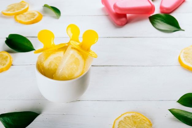 Lemon popsicles in a cup on a white wooden table decorated with lemon slices and leaves from nature