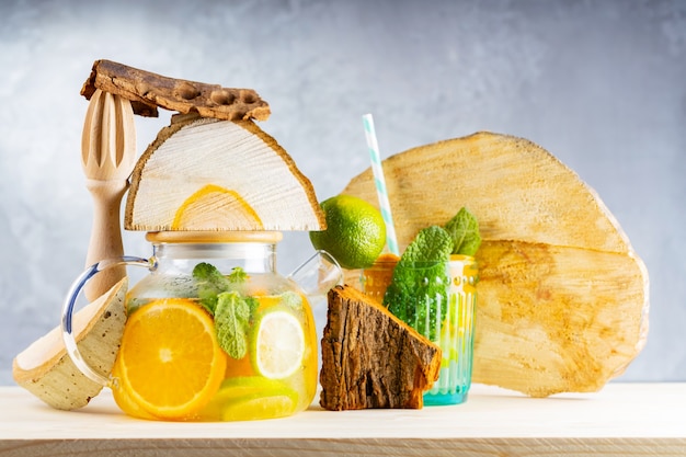 Lemonade drink of water, lemon, orange and mint leaves in a transparent teapot. Lime mint iced tea and pieces of wood. Creative composition