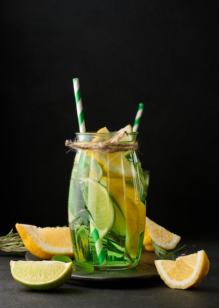 Lemonade in a transparent glass with lemon lime rosemary sprigs and mint leaves on a black background