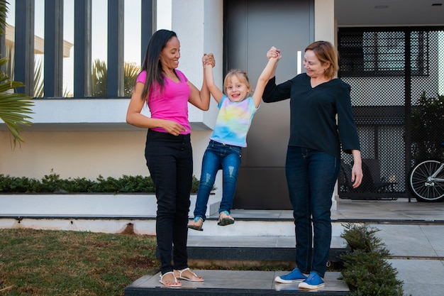 Lesbian couple holding foster daughter's hand at home door. Lesbian couple with daughter. Adoption and diversity concept