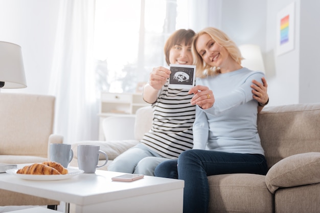 Lesbian mothers. Happy nice female couple sitting together and showing you a photo while expecting a baby