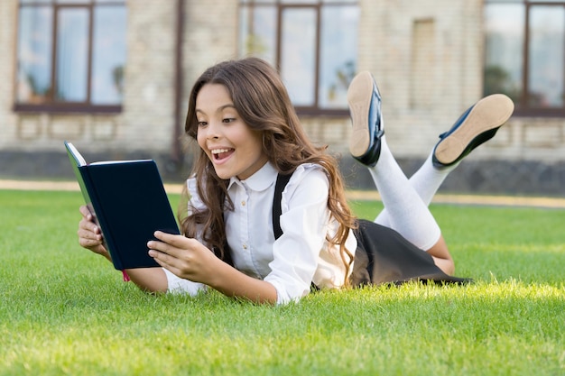 Lesson on fresh air Cute small child reading book outdoors Educational book Literature for kids Schoolgirl school uniform laying on lawn with favorite book Bookworm concept Access to knowledge