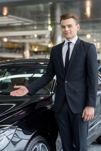 Let me present our best model. Handsome young car salesman standing at the dealership and pointing car