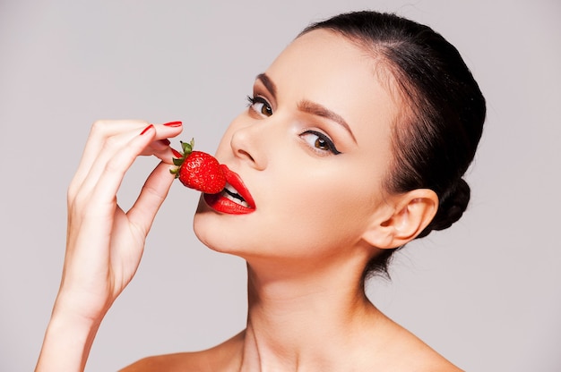 Let me tempt you! Beautiful young shirtless woman holding strawberry in her hand while standing against grey background