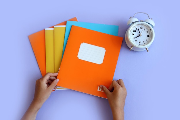 Let's go back to school the child's hands hold an orange notepad next to a white alarm clock purple background top view space for text