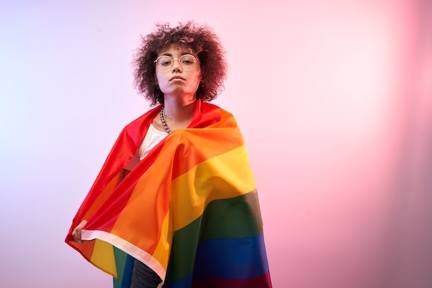 Lgbtq concept. Positive caucasian girl with afro curly hair holding rainbow flag isolated in studio