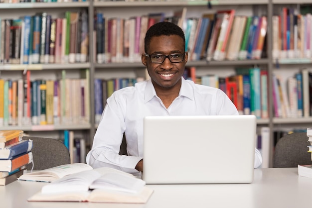 In The Library  Handsome African Male Student With Laptop And Books Working In A High School  University Library  Shallow Depth Of Field