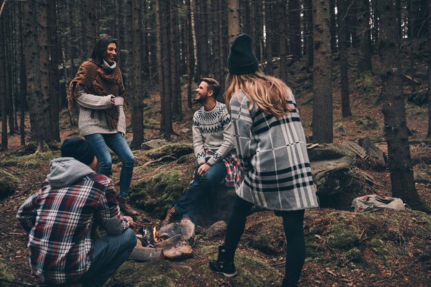 Life filled with friendship. Group of happy young people standing around the campfire while hiking in the woods