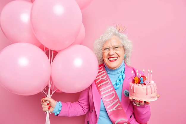 Life only start when you get older. Glad wrinkled old woman celebrates birthday holds festive cake with burning candled bunch of inflated balloons