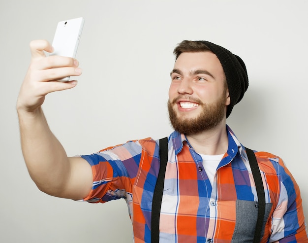 Life style concept: a young man with a beard  in shirt holding mobile phone and making photo of himself while standing against grey background.