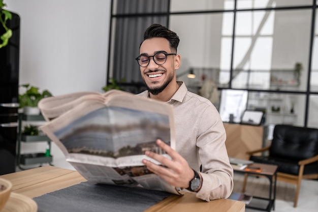 Life style. Happy attractive successful man in glasses smiling reading newspaper at home sitting at table