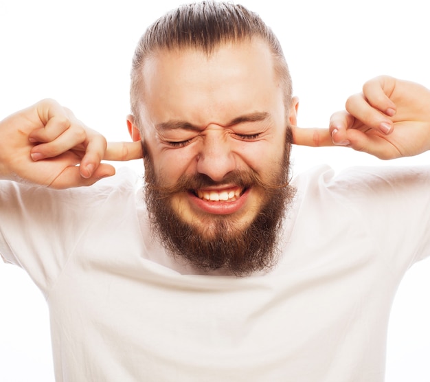 Life style and people concept: frustrated bearded man in white shirt holding fingers in his ears and keeping eyes closed while standing against white background